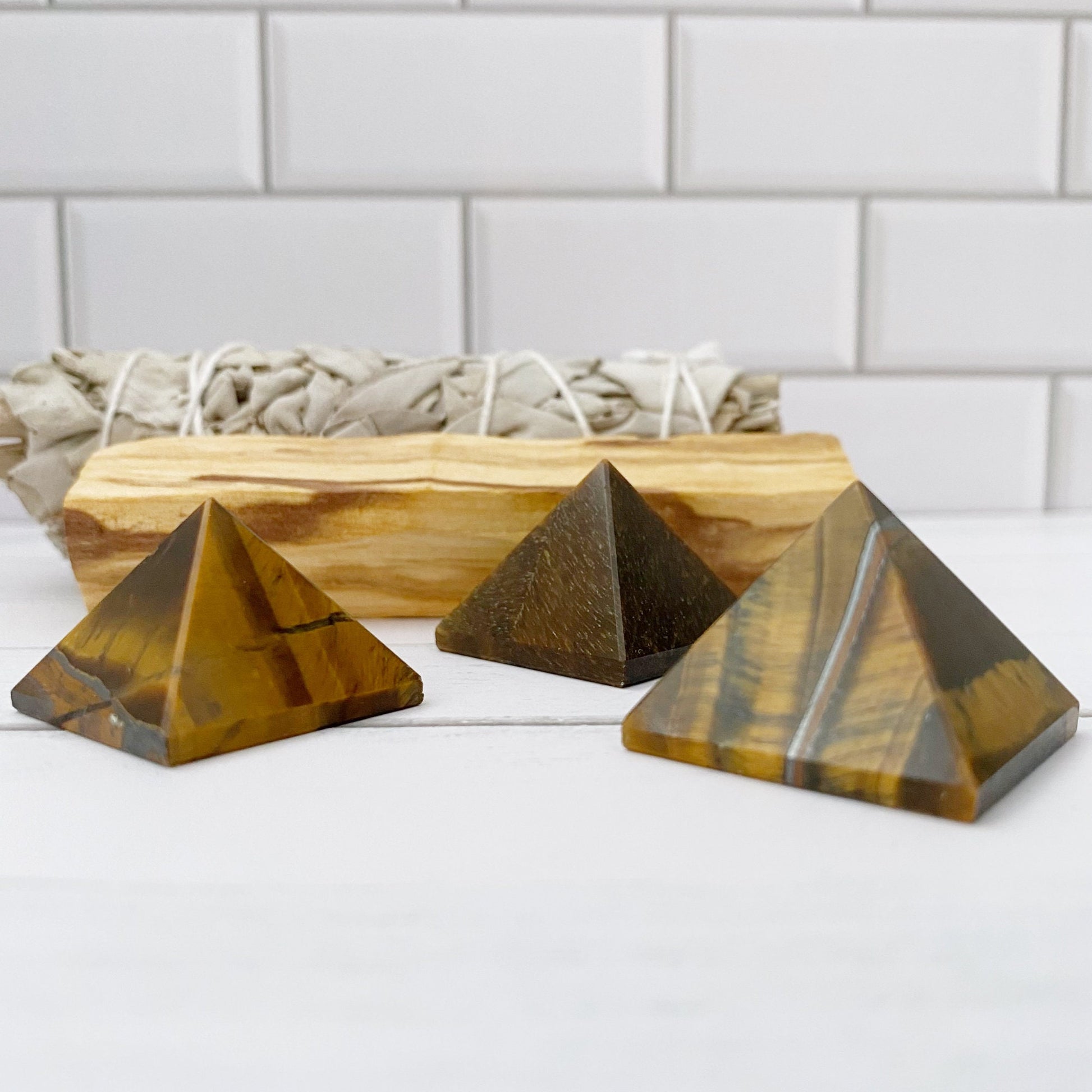 Three small 1" Mini Tigers Eye Pyramids are displayed on a white surface against a white subway tile background. Behind the pyramids, there is a bundle of sage and a piece of Palo Santo wood, enhancing their aura of spiritual awareness.