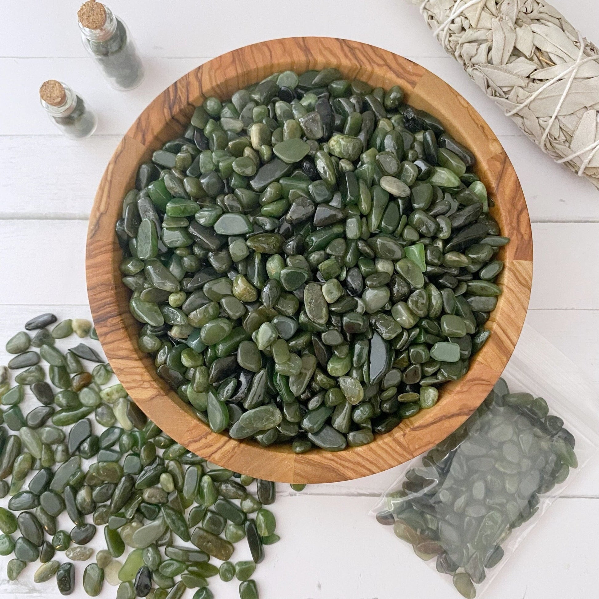 A wooden bowl filled with polished Nephrite Jade Crystal Chips sits on a white surface. More tumbled and polished stones are scattered next to the bowl, while a plastic bag holds additional ones nearby. In the background, there’s a white sage bundle and small bottles, perfect for creative and mystical endeavors.