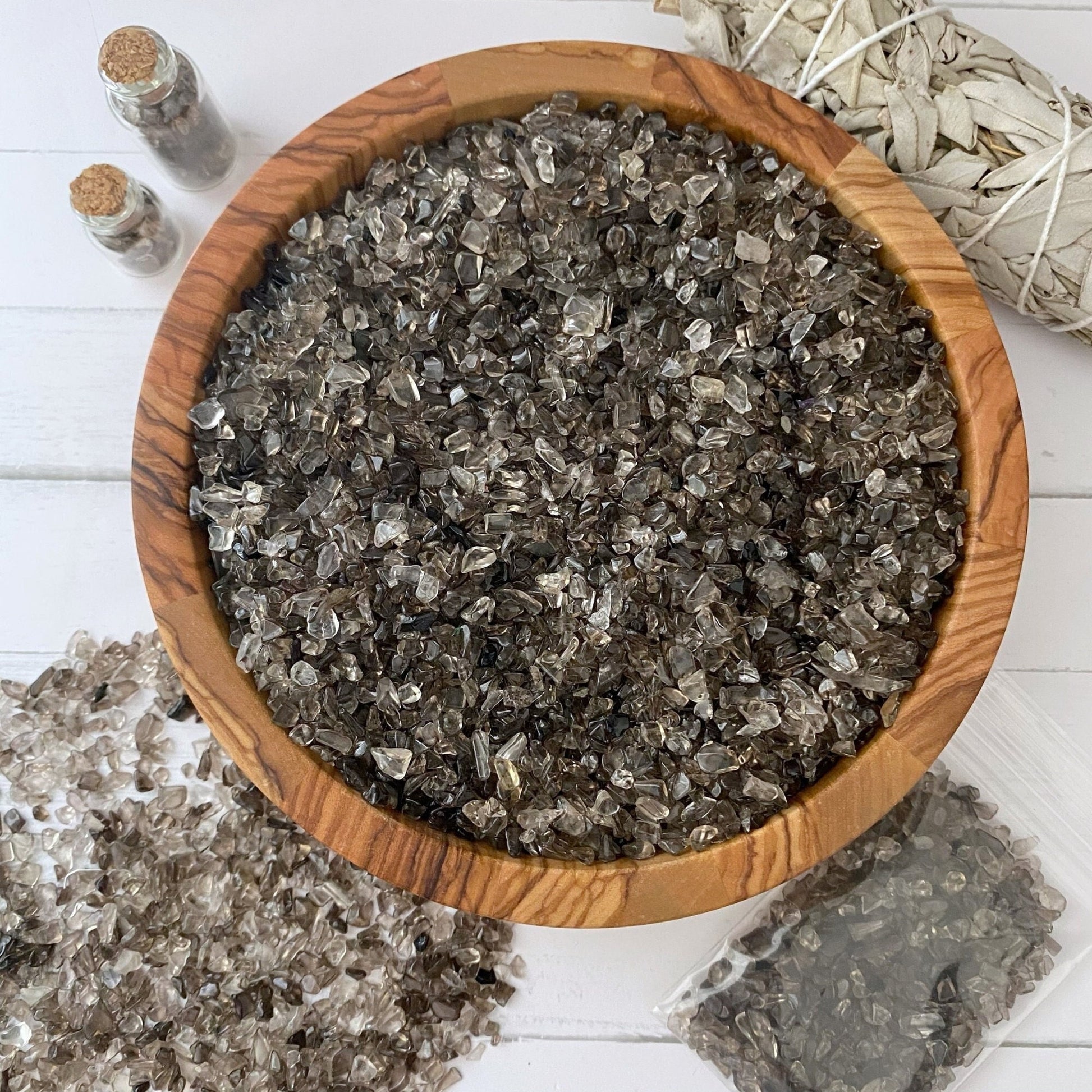 A wooden bowl filled with small, rough, smoky quartz crystal chips sits on a white surface. Adjacent to the bowl are several loose crystals, small vials of crystals with cork stoppers, a piece of white sage, and a small plastic bag of additional crystals for your mystical endeavors.