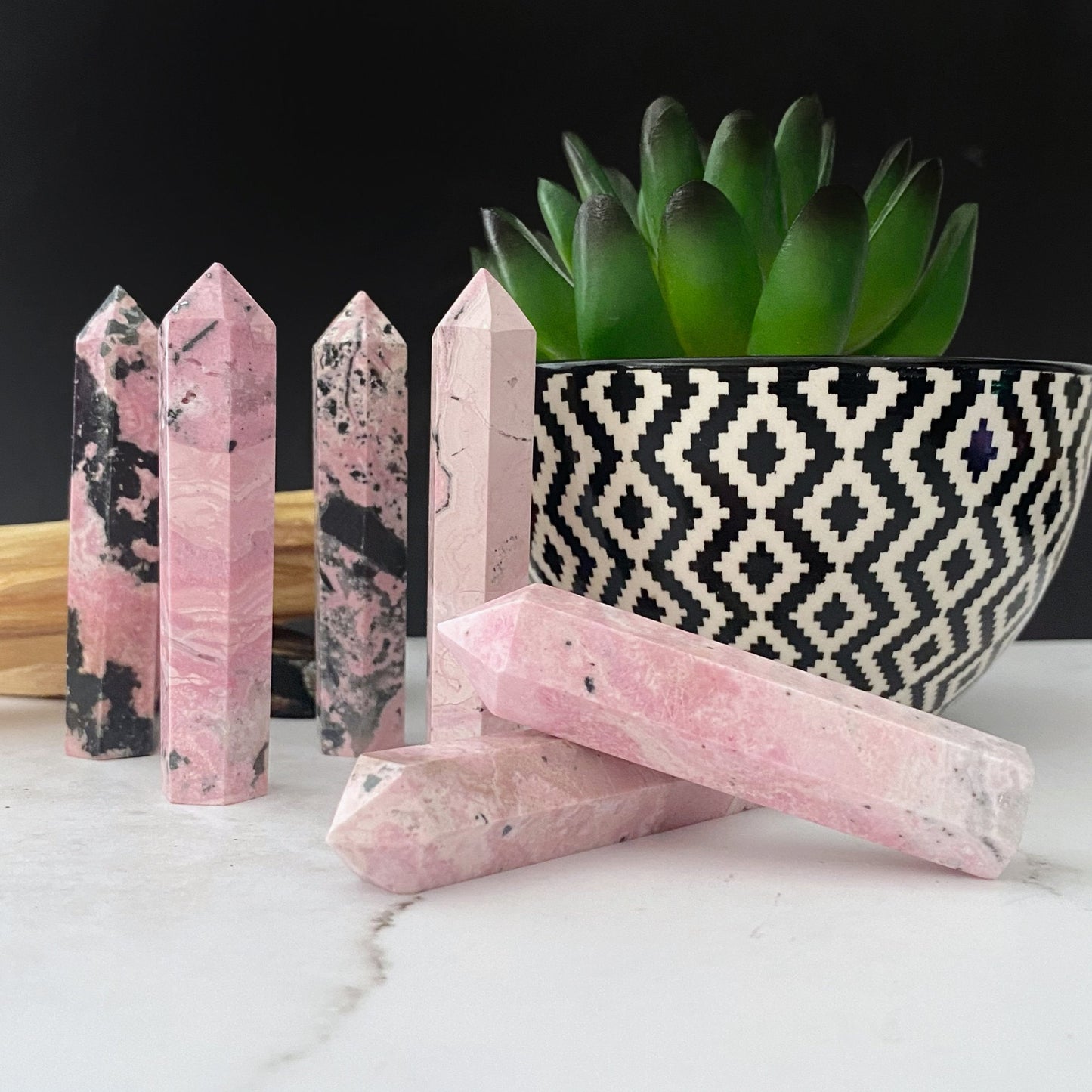 Several pink crystal towers, including rhodochrosite and a 2-3" Rhodonite Point, stand upright on a white surface with a potted succulent in a black and white geometric-patterned pot in the background. A wooden piece is also visible beside the crystals, creating an enchanting crystal grid for heart chakra alignment.