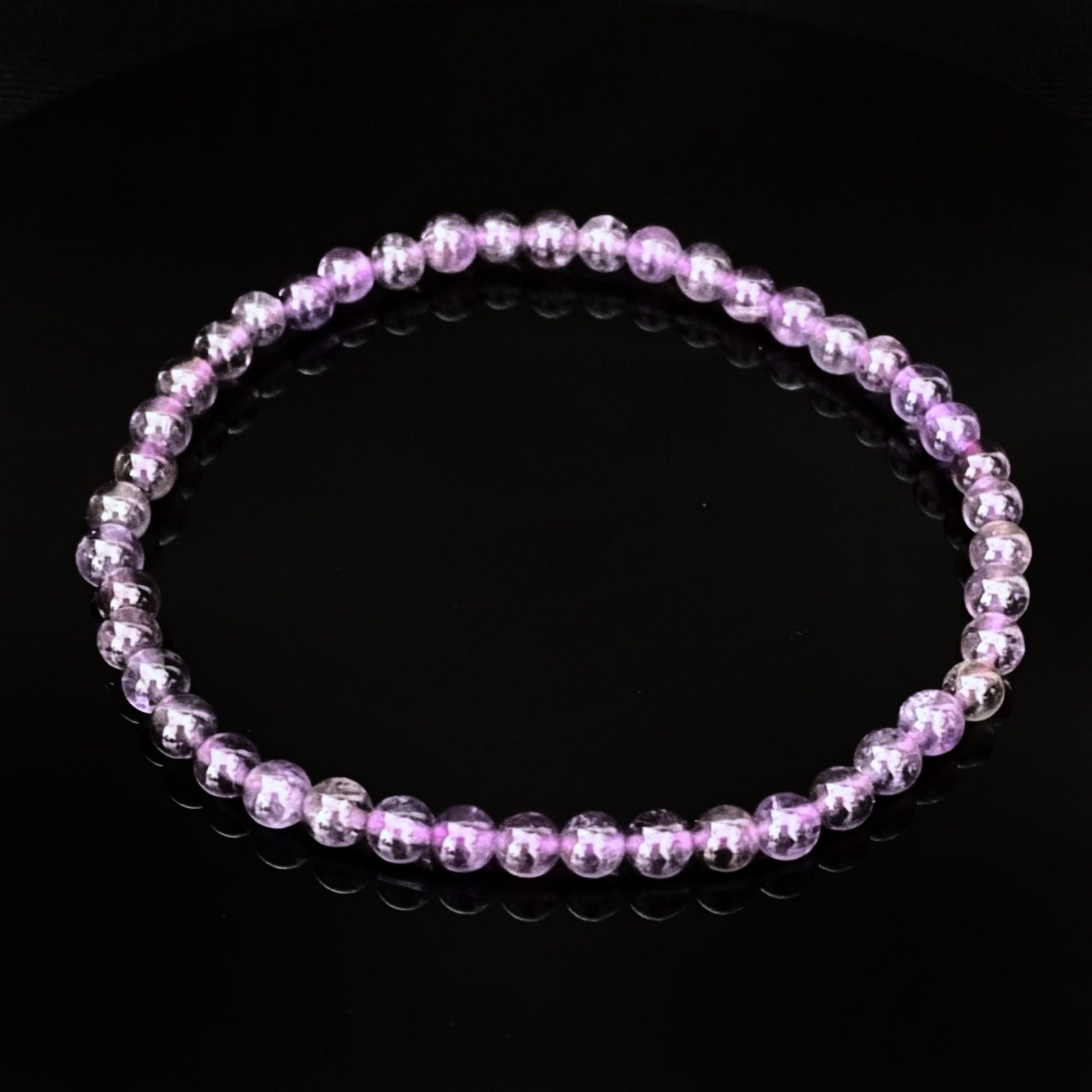 A close-up of a 4mm Amethyst Beaded Bracelet consisting of small, round, translucent purple beads, arranged in a circular pattern and placed against a dark background. The reflection of the bracelet is visible on the glossy surface below it.