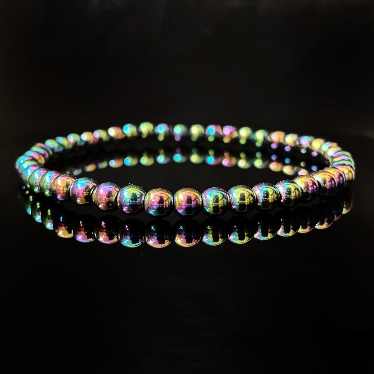 A close-up of a 4mm Rainbow Hematite Beaded Bracelet composed of iridescent, round Rainbow Hematite beads. The bracelet forms a perfect circle and is placed against a shiny black background, creating a mirrored reflection underneath the beads.