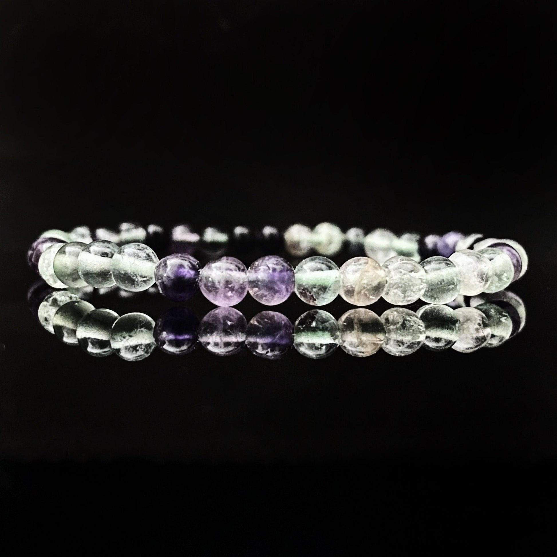 A 4mm Rainbow Fluorite Beaded Bracelet with spherical, translucent beads in various shades of purple, green, and clear rests on a shiny black surface. The surface reflects the bracelet, creating a mirror-like effect while symbolizing boosted creativity & clarity through its energetic cleansing properties.