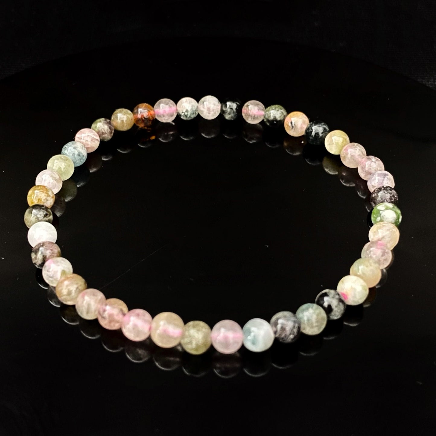 A delicate beaded 4mm Rainbow Tourmaline Bracelet with spherical beads in various shades of pastel colors, including pink, green, and blue, lies on a reflective black surface. The reflection of the bracelet is clearly visible, creating a symmetrical effect and enhancing its chakra balancing properties.