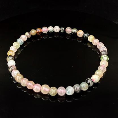 A delicate beaded 4mm Rainbow Tourmaline Bracelet with spherical beads in various shades of pastel colors, including pink, green, and blue, lies on a reflective black surface. The reflection of the bracelet is clearly visible, creating a symmetrical effect and enhancing its chakra balancing properties.