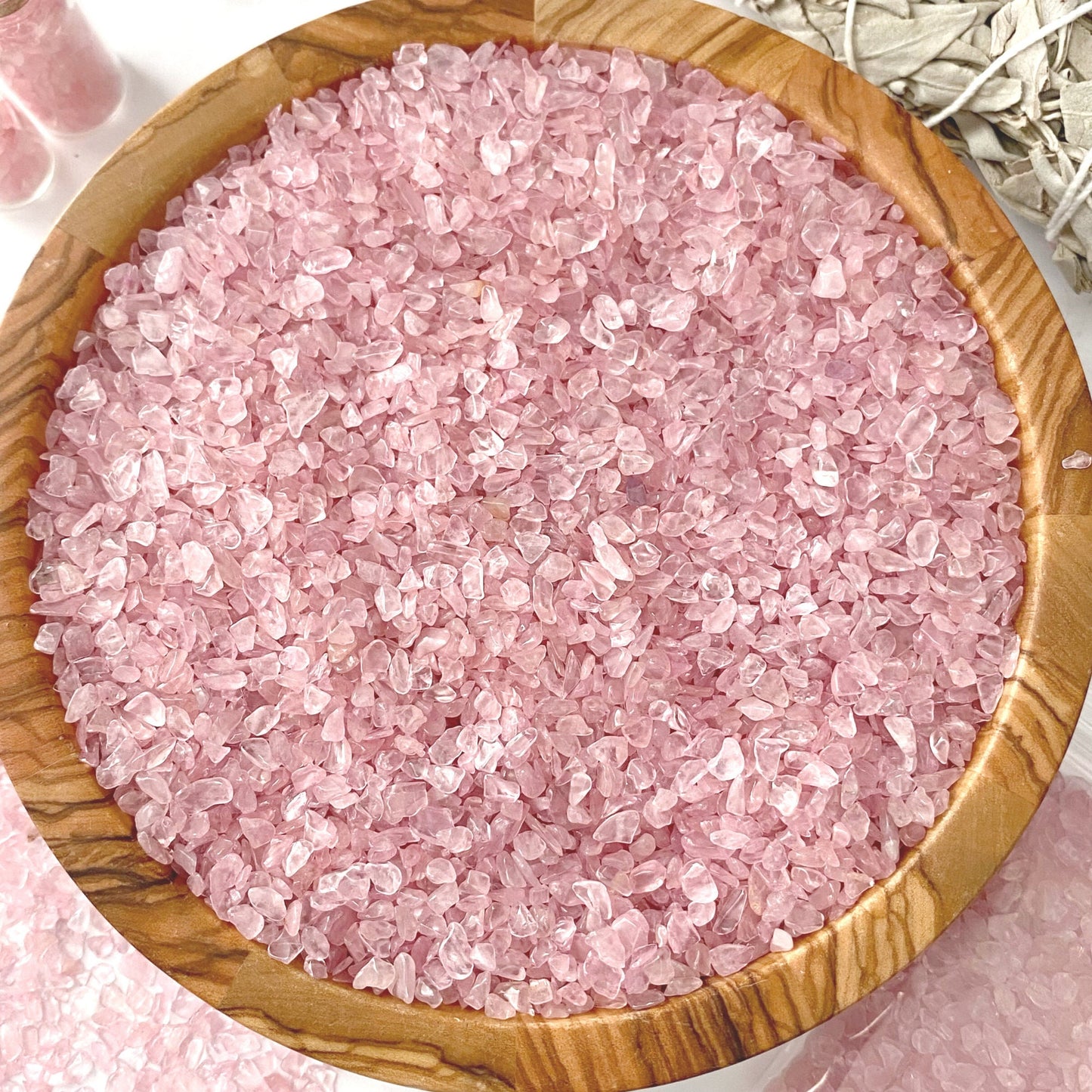 A wooden bowl filled with Rose Quartz Crystal Chips sits on a marble surface. Nearby, two small glass vials with pink salt and a plastic bag containing more crystals accompany a bundle of dried sage wrapped with string. Some pink salt is scattered, mingling gracefully with Rose Quartz Crystal Chips on the surface.