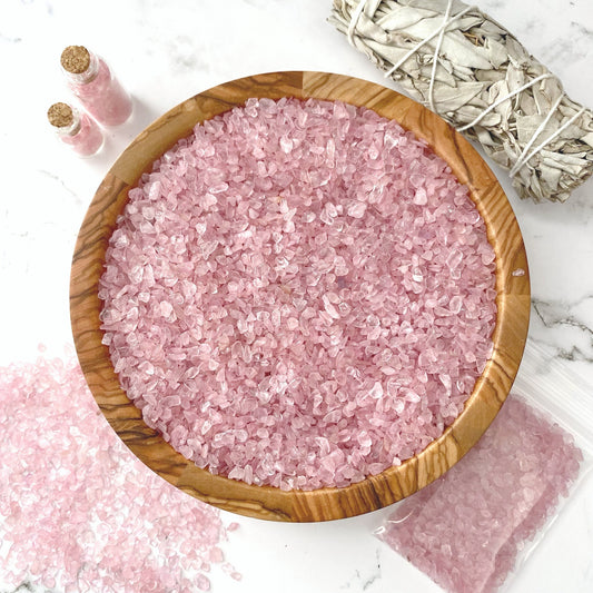 A wooden bowl filled with Rose Quartz Crystal Chips sits on a marble surface. Nearby, two small glass vials with pink salt and a plastic bag containing more crystals accompany a bundle of dried sage wrapped with string. Some pink salt is scattered, mingling gracefully with Rose Quartz Crystal Chips on the surface.