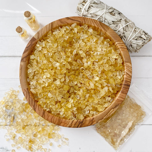 A wooden bowl filled with small, yellow Citrine Crystal Chips sits on a white surface. Surrounding the bowl are a small bundle of sage, two tiny glass bottles filled with similar crystals, and a small plastic bag containing additional stones.