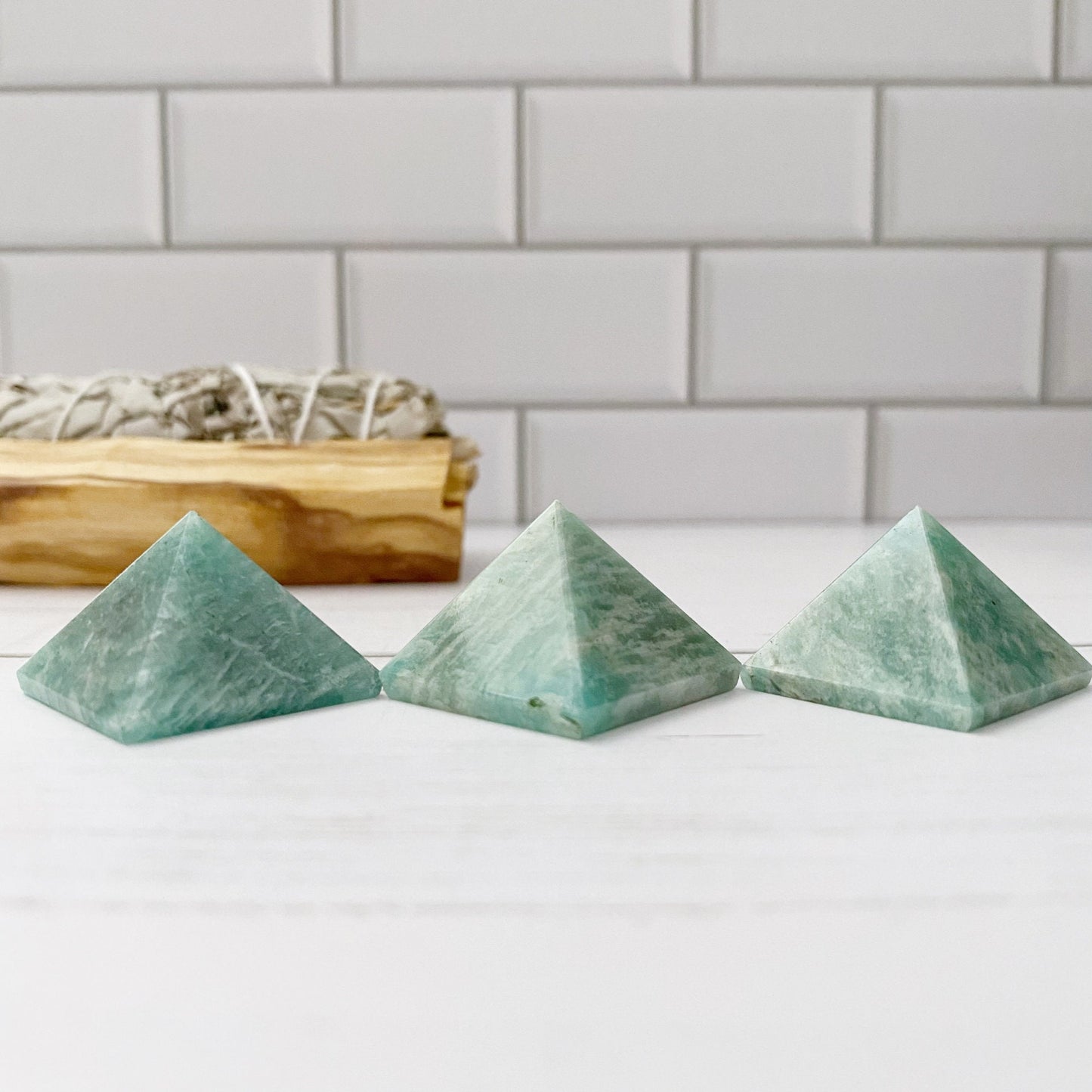 A photo features three 1" Mini Amazonite Pyramids on a white surface, known for stress relief and combating electromagnetic pollution. Behind the pyramids, there is a bundle of dry, white sage sticks. The background consists of white tiled walls, adding a clean and minimalist look to the setup.