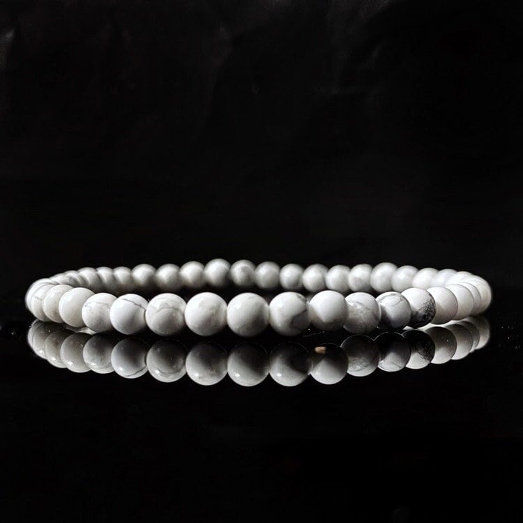 A 4mm Howlite Beaded Bracelet made of white marble-like stones with subtle gray veining is displayed against a black reflective surface, highlighting the bracelet's elegant and smooth texture. It’s perfect for those seeking emotional healing and balance through Usui Reiki energy.