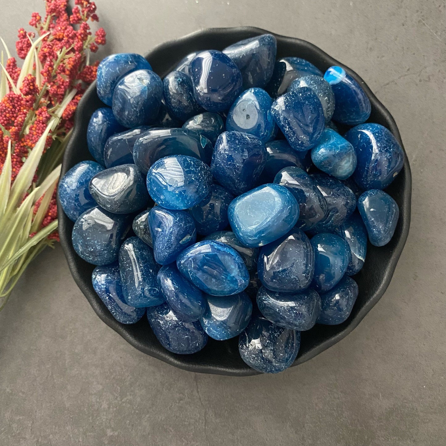 A black bowl filled with polished, shiny Blue Onyx Tumbled Stones is placed on a dark gray surface. To the left of the bowl, there is a sprig of red and green flowers.