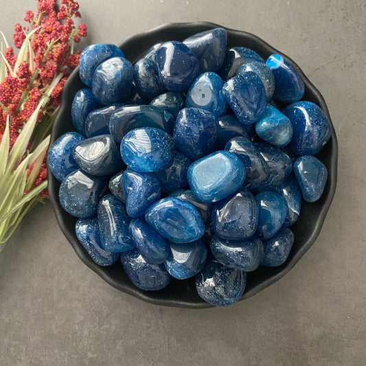 A black bowl filled with polished, shiny Blue Onyx Tumbled Stones is placed on a dark gray surface. To the left of the bowl, there is a sprig of red and green flowers.