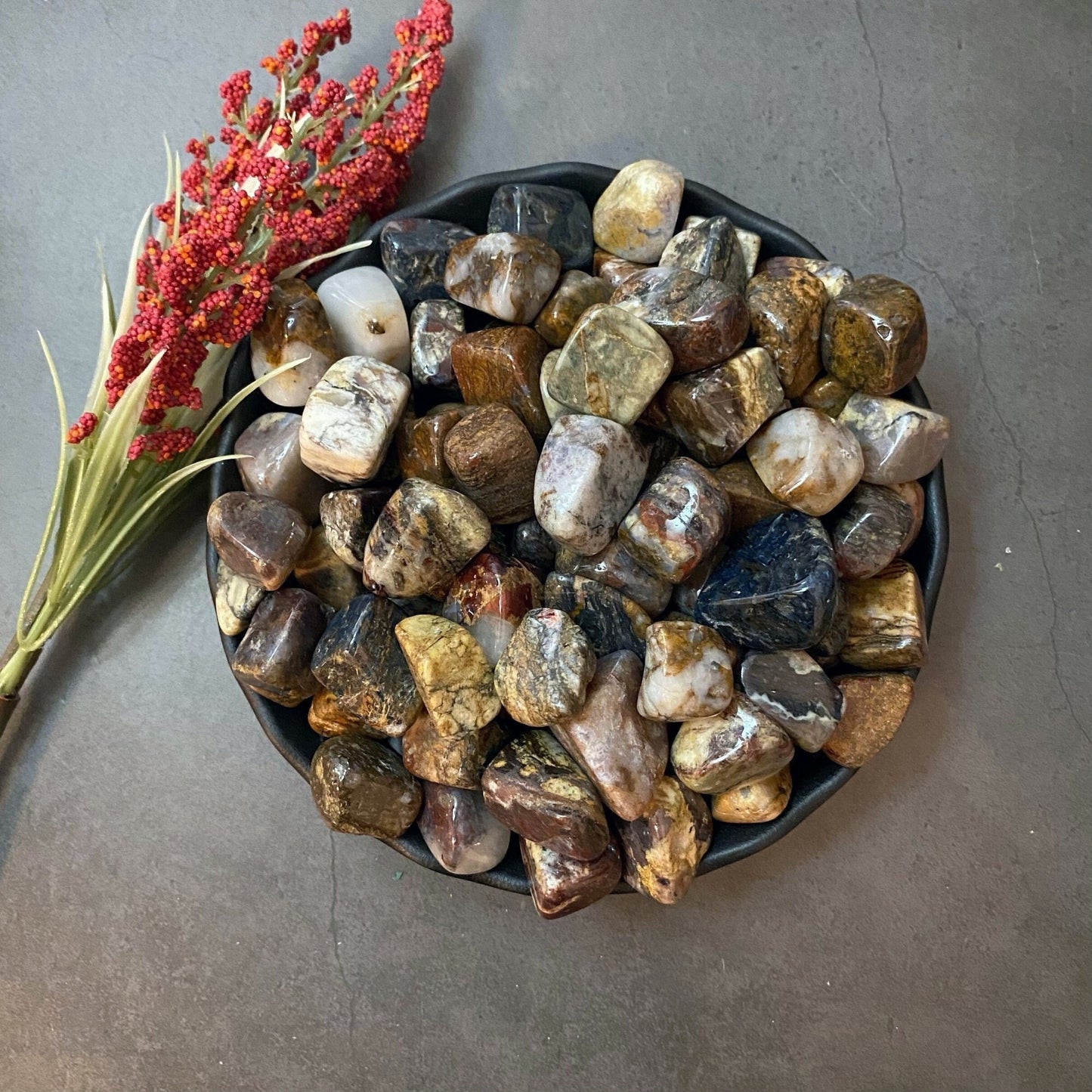 A black plate filled with polished stones of various earthy colors, including browns, grays, and blues, is placed on a gray surface. Among them are Pietersite Tumbled Stones known for their connection to the third eye chakra. A small branch with red berries and green leaves lies next to the plate.