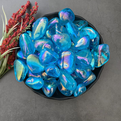 A black bowl is filled with blue iridescent glass pebbles and Aqua Aura Quartz Tumbled Stones, their glossy surfaces reflecting rainbow colors. The bowl is placed on a dark grey countertop, with a sprig of red and green foliage next to it, creating a serene space perfect for Throat Chakra healing.