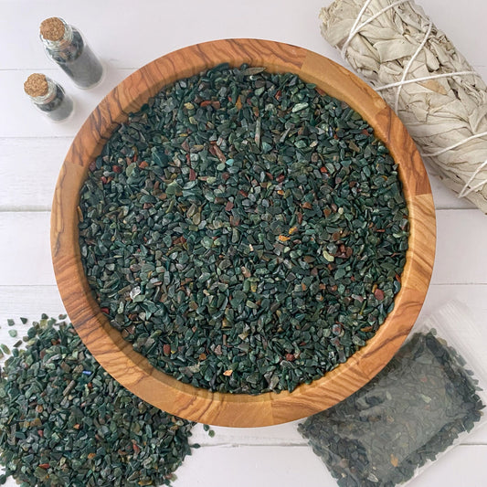 A wooden bowl filled with small, polished green healing stones is placed on a white surface. Next to the bowl are a bundle of white sage, two tiny glass bottles, and a plastic bag containing more stones. Some Bloodstone Crystal Chips are scattered around, radiating natural energy.