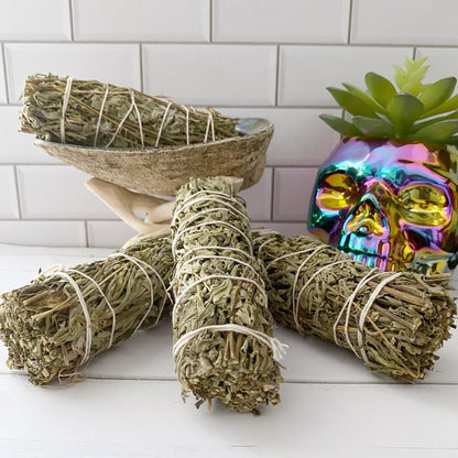 A display of sage and Organic Mugwort Smudge Sticks wrapped in white string is placed in a white room with a tiled wall. Some bundles are inside a seashell, supported by a stand. A colorful, metallic skull decor and a succulent plant sit beside the sage bundles, creating an ideal setting for smudging rituals.