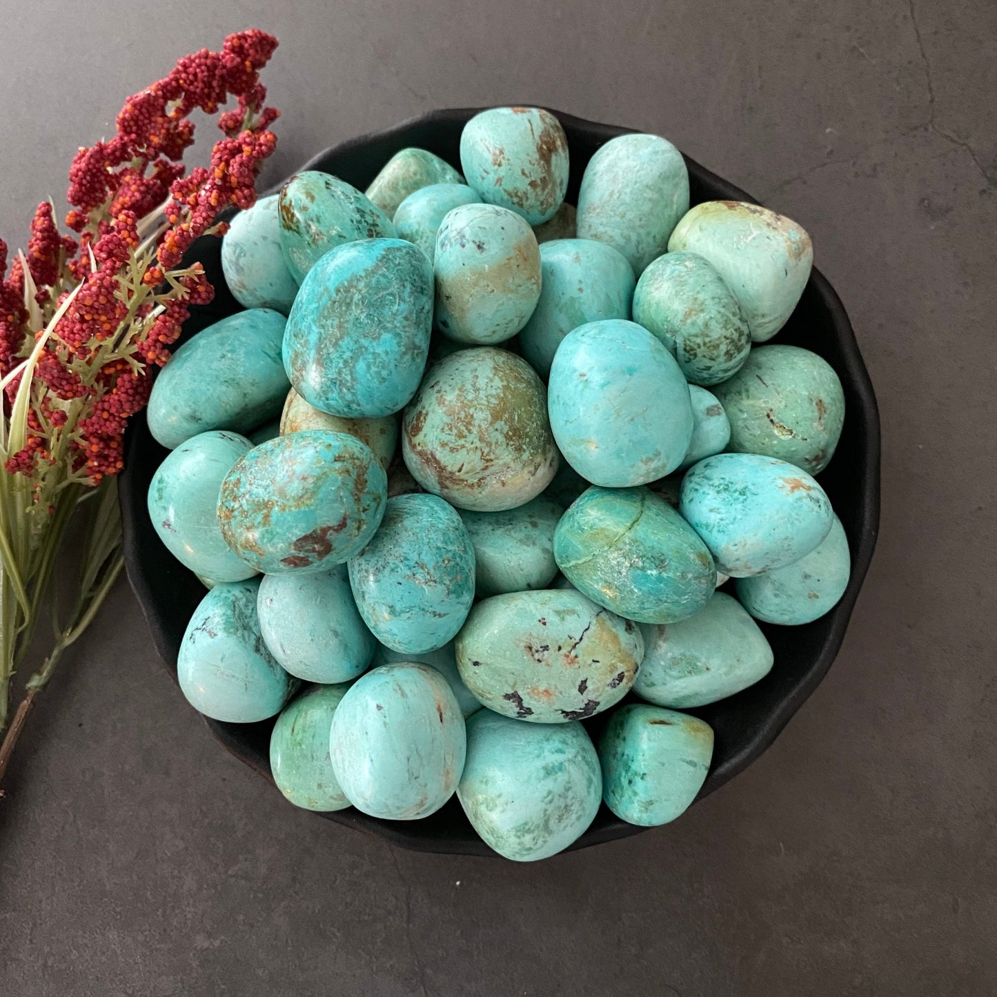 A black bowl filled with numerous Peruvian Turquoise Tumbled Stones, exhibiting natural variations in color and pattern. To the left of the bowl, a bunch of red dried flowers rests on a dark, textured surface, perfect for chakra balancing.