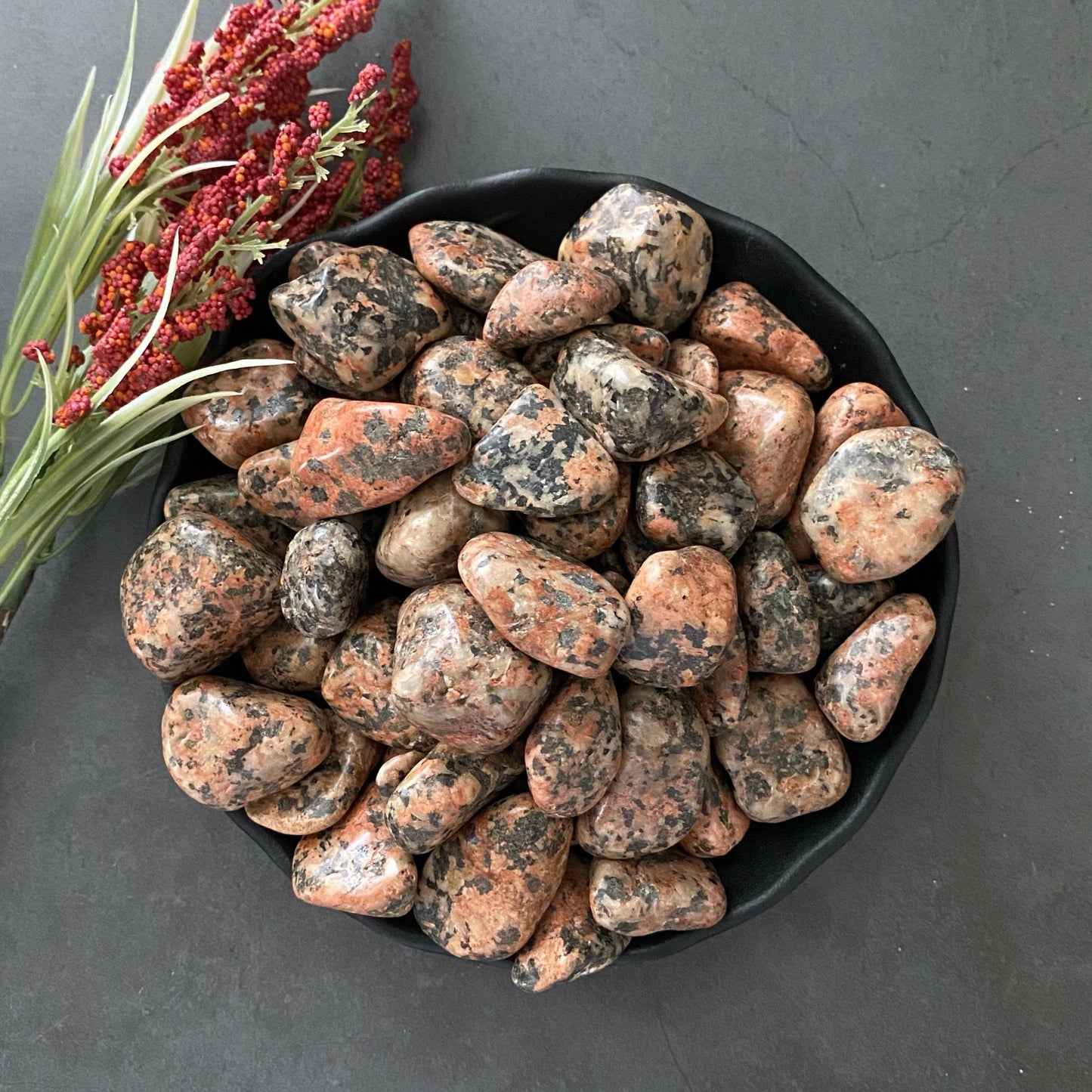 A black bowl filled with polished, multicolored stones, including soothing Orthoclase Tumbled Stones, varying in shades of pink, gray, and black sits on a dark surface. To the left of the bowl, a small bouquet of red and green dried flowers adds a natural touch to the scene. Perfect for those looking to align chakras.