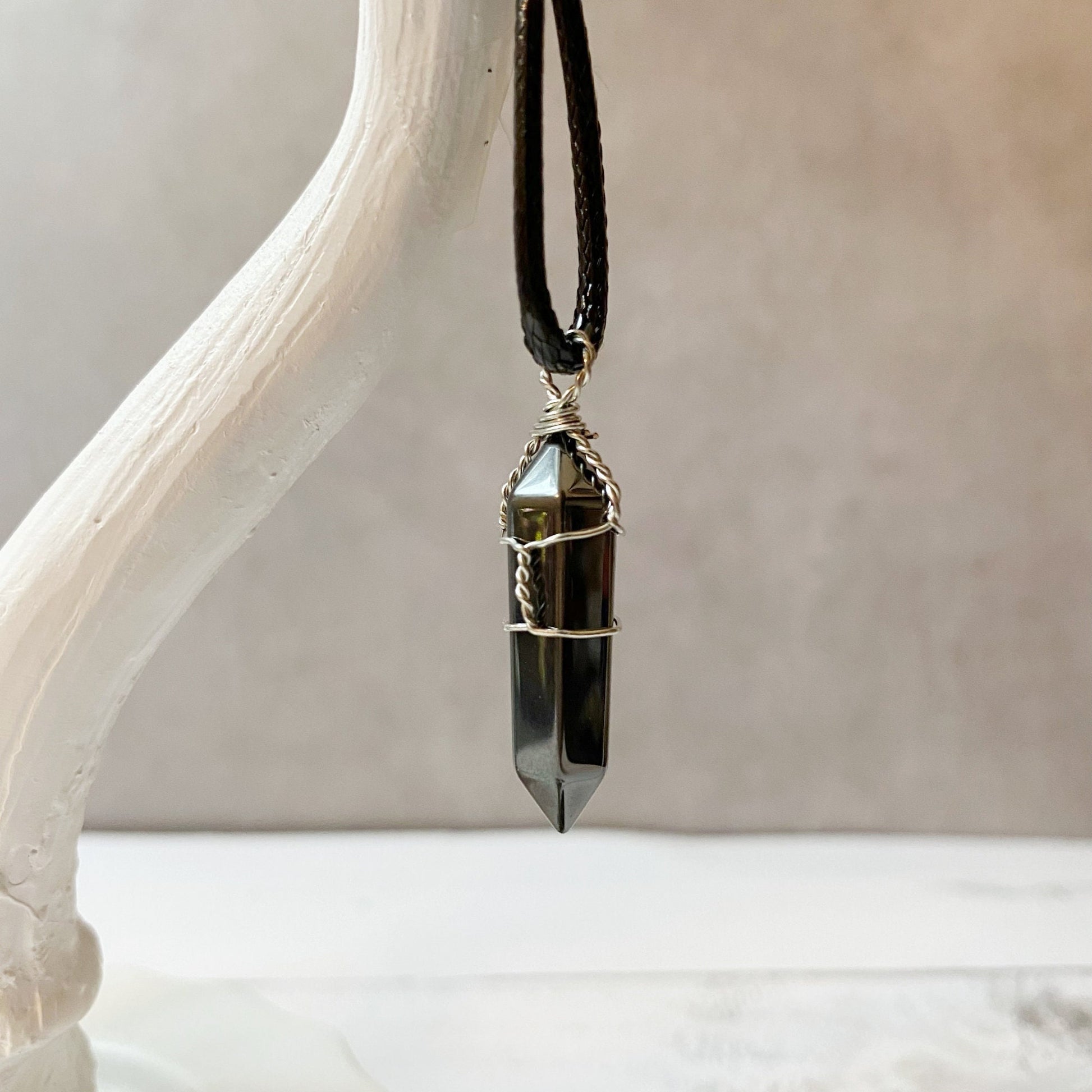 A polished, dark crystal pendant wrapped with silver wire is suspended from a black braided cord necklace. The background is blurred to highlight the 1.5" Double Terminated Hematite Point Pendant, which has a pointed, hexagonal shape. This wire-wrapped jewelry piece exudes minimal and elegant charm while offering grounding energy.