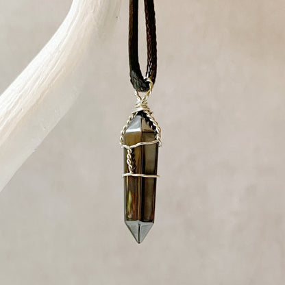 A polished, dark crystal pendant wrapped with silver wire is suspended from a black braided cord necklace. The background is blurred to highlight the 1.5" Double Terminated Hematite Point Pendant, which has a pointed, hexagonal shape. This wire-wrapped jewelry piece exudes minimal and elegant charm while offering grounding energy.