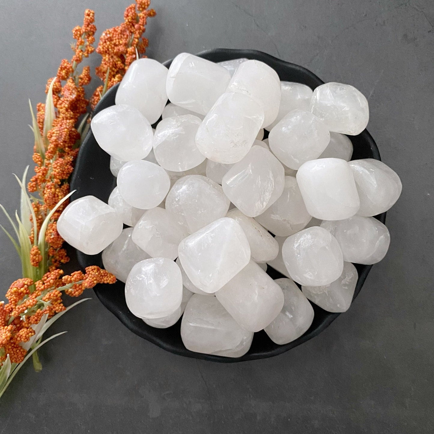 A black bowl filled with smooth, white, rounded Phantom Quartz Tumbled Stones is placed on a dark surface. To the left of the bowl is a sprig of orange and green flora, adding a natural touch to the arrangement. This serene setting invites meditation and healing, fostering spiritual growth through its simple beauty.