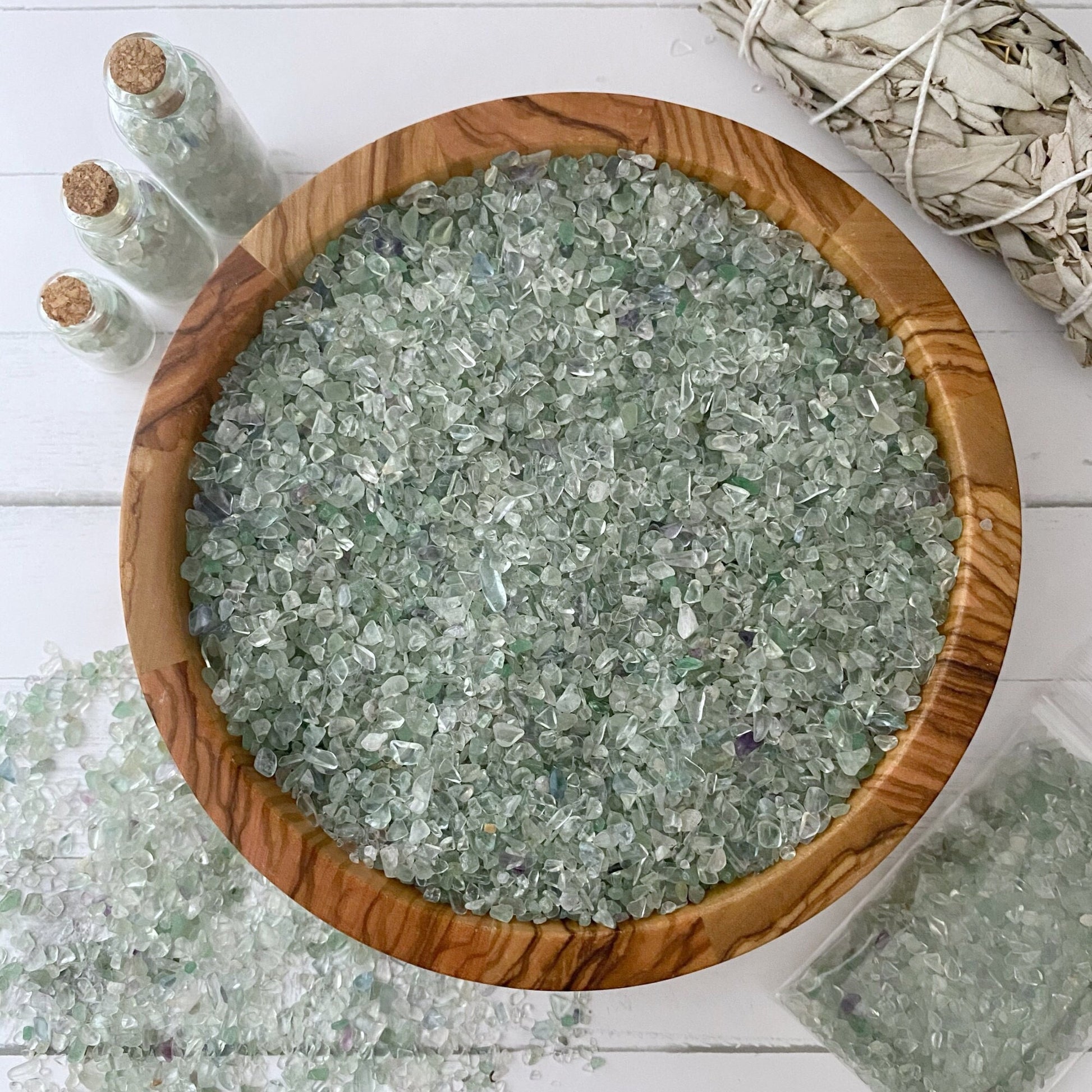 A wooden bowl filled with small, translucent Green Fluorite Crystal Chips is centered on a white surface. Nearby, three small glass bottles also contain Green Fluorite Crystal Chips, and there is a bundle of dried sage in the upper right corner. Some tumbled and polished stones are spilled beside the bowl.