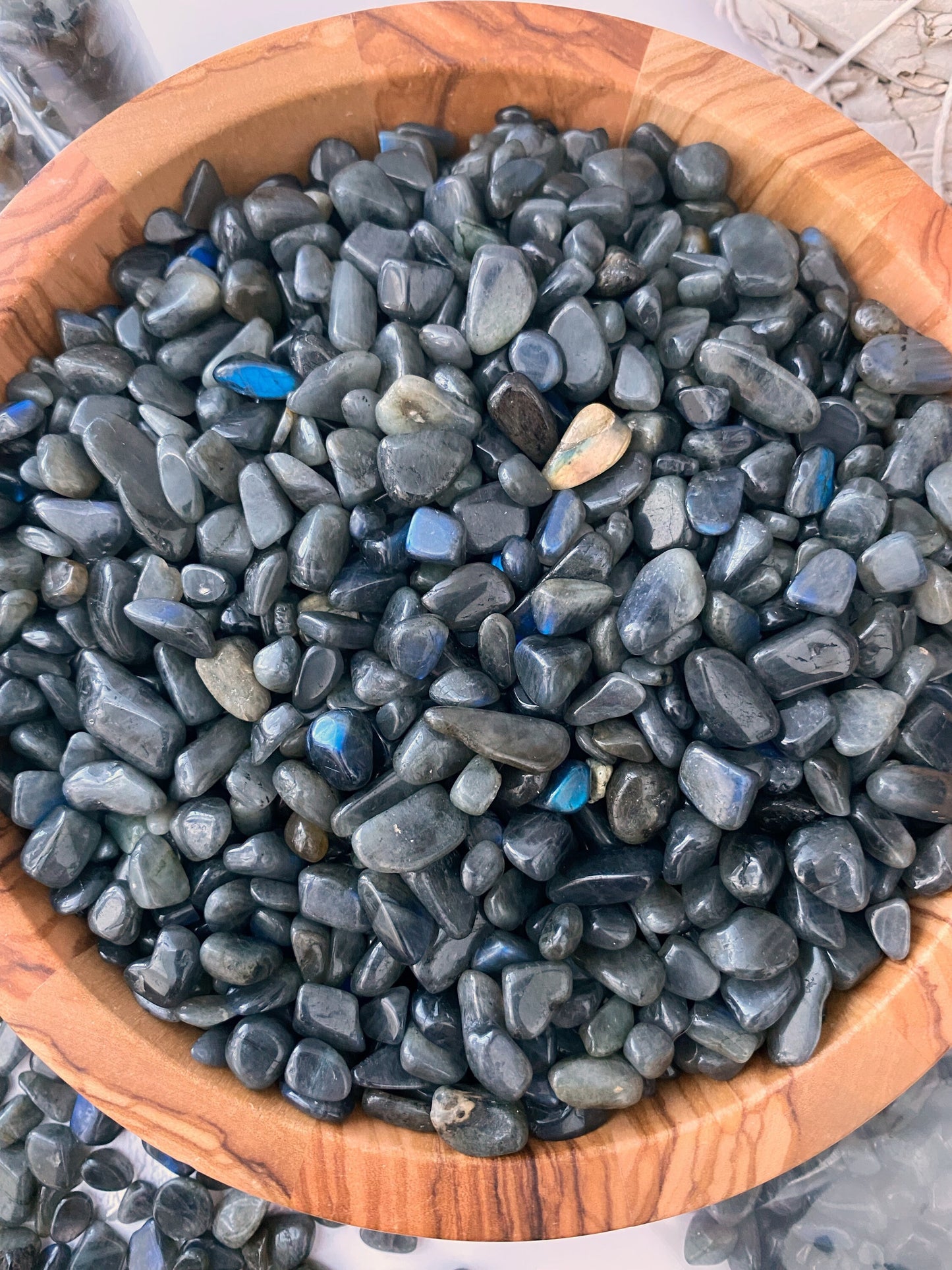 A wooden bowl filled with polished black stones is placed on a white surface. Surrounding the bowl are small glass bottles with cork stoppers, a bundle of white sage, and more black stones scattered around and in a plastic bag. Among the items, Labradorite Crystal Chips shimmer subtly, adding to the mystic allure.