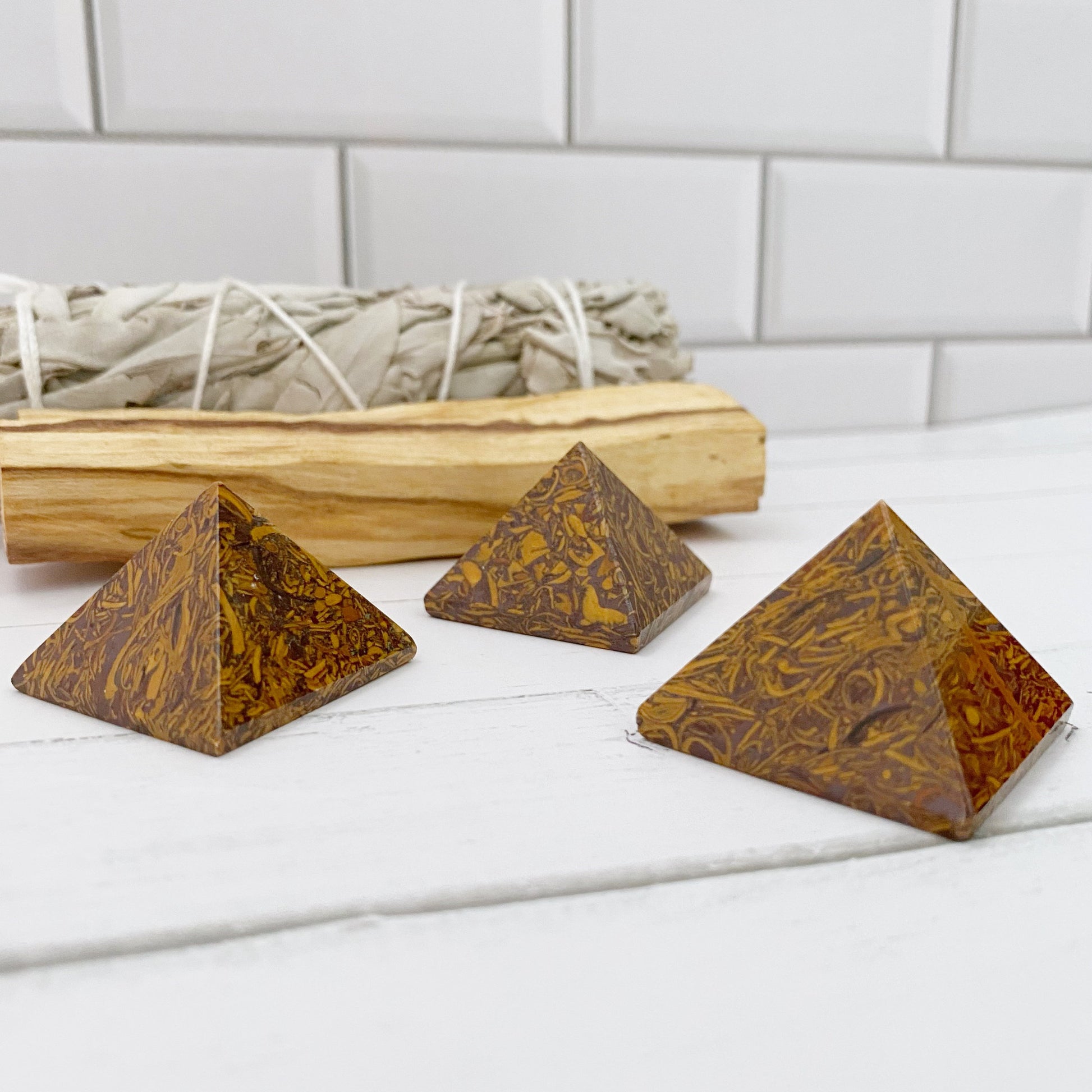 Three small, brown 1" Mini Calligraphy Jasper Pyramids with intricate patterns sit on a white surface in front of a white brick wall. Behind the pyramids, there is a bundle of sage tied with white string, resting on a wooden tray.