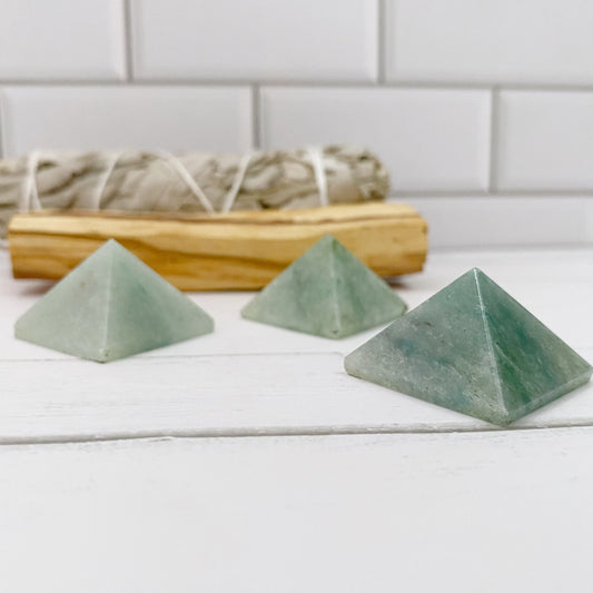 Three 1" Mini Green Aventurine Pyramids are arranged on a white wooden surface with a bundle of sage in the background. Perfect for meditation, the scene is set against white tiled walls.