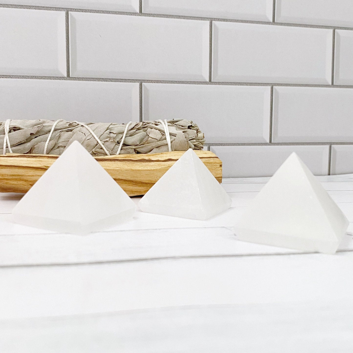 Three polished 1.5" Mini Selenite Pyramids are arranged on a white surface in front of a backdrop of white subway tiles. Behind the pyramids, embodying sacred geometry, is a bundle of dried sage tied with string, resting on a piece of wood. The setting appears calm and minimalistic, perfect for mental clarity.