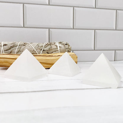 Three polished 1.5" Mini Selenite Pyramids are arranged on a white surface in front of a backdrop of white subway tiles. Behind the pyramids, embodying sacred geometry, is a bundle of dried sage tied with string, resting on a piece of wood. The setting appears calm and minimalistic, perfect for mental clarity.