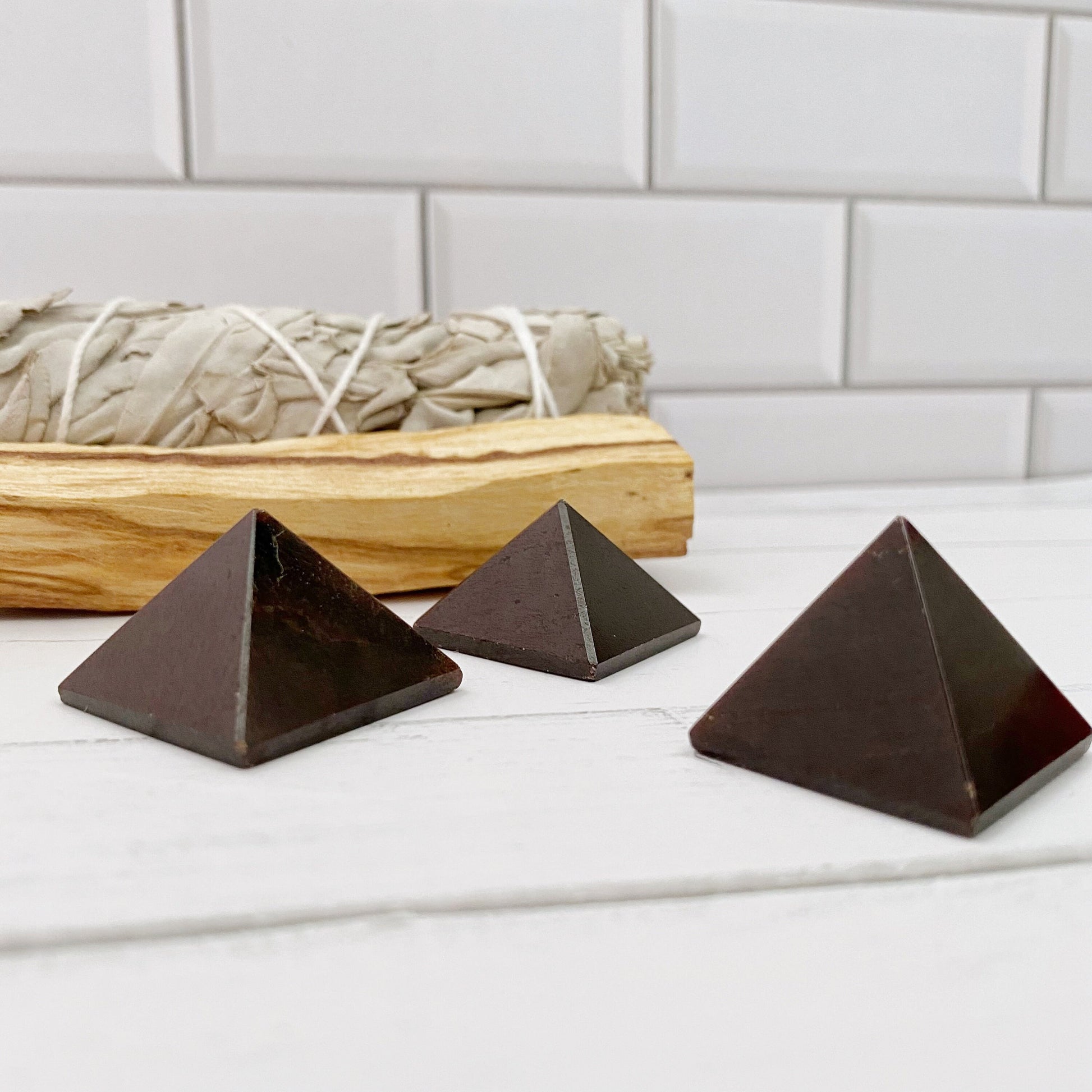 Three 1" Mini Garnet Pyramids, likely made of stone or crystal, are placed in front of a bundle of sage tied with twine. The scene is set against a white tiled background. The objects and sage are on a light-colored surface, creating a minimalist aesthetic perfect for chakra cleansing and emotional balance.