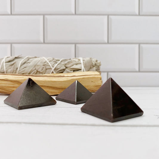 Three 1" Mini Garnet Pyramids, likely made of stone or crystal, are placed in front of a bundle of sage tied with twine. The scene is set against a white tiled background. The objects and sage are on a light-colored surface, creating a minimalist aesthetic perfect for chakra cleansing and emotional balance.