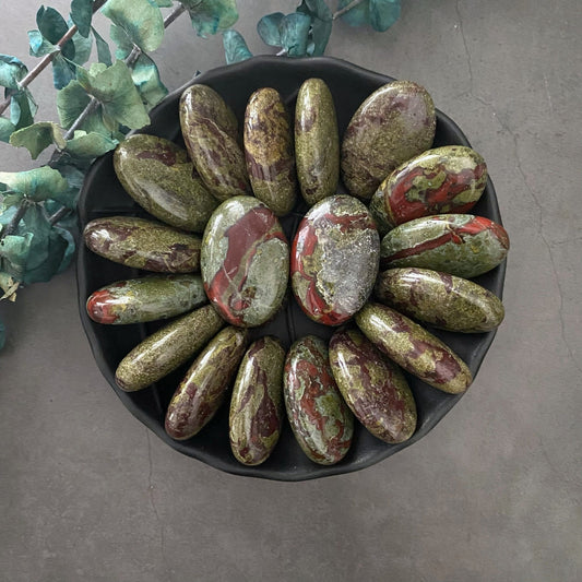 A black bowl filled with polished, oval-shaped stones featuring a mix of green, red, and brown hues, including Dragon Stone Palm Stone for inner strength. These stones are arranged in a flower-like pattern. The bowl sits on a gray surface adorned with green foliage around it.