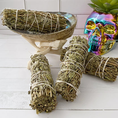 A display of sage and Organic Mugwort Smudge Sticks wrapped in white string is placed in a white room with a tiled wall. Some bundles are inside a seashell, supported by a stand. A colorful, metallic skull decor and a succulent plant sit beside the sage bundles, creating an ideal setting for smudging rituals.