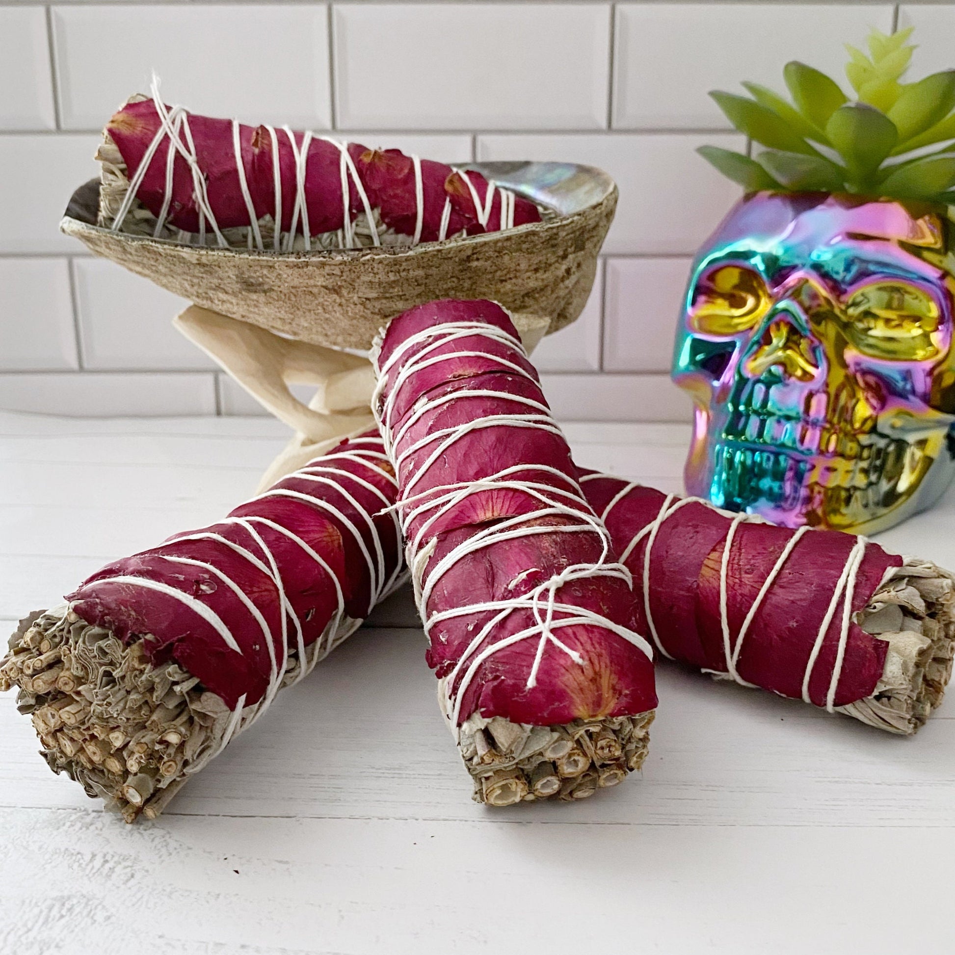 An Organic White Sage & Red Rose Petals bundle wrapped with white string is displayed in an abalone shell on a stand. Nearby, a colorful, reflective skull and a succulent plant add charm against the white tile background, making for an elegant energy cleansing setup.