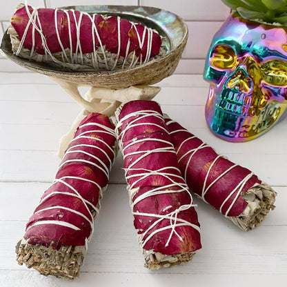 An Organic White Sage & Red Rose Petals bundle wrapped with white string is displayed in an abalone shell on a stand. Nearby, a colorful, reflective skull and a succulent plant add charm against the white tile background, making for an elegant energy cleansing setup.