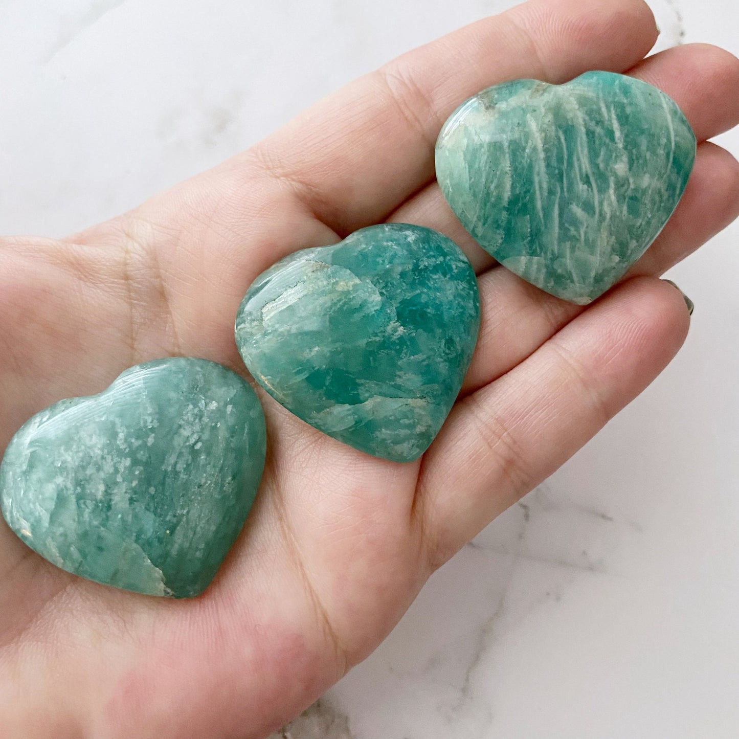 Ten heart-shaped 1.5" Small Amazonite Crystal Hearts on a white marble surface, arranged in a circular pattern. A bundle of white sage is seen on the upper left and a colorful, iridescent skull-shaped container is on the upper right.