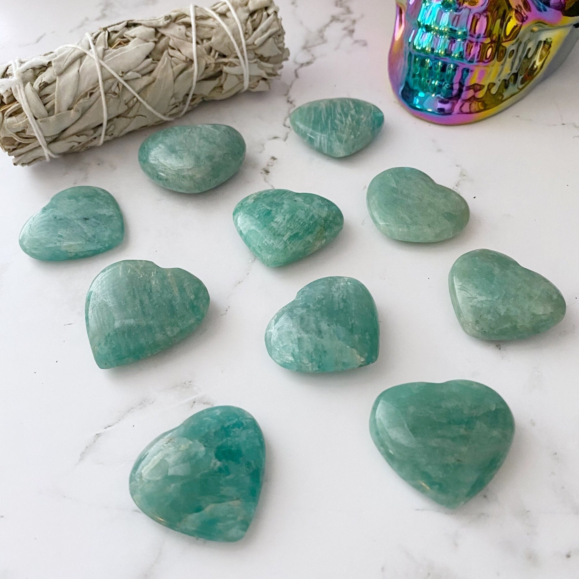 Ten heart-shaped 1.5" Small Amazonite Crystal Hearts on a white marble surface, arranged in a circular pattern. A bundle of white sage is seen on the upper left and a colorful, iridescent skull-shaped container is on the upper right.