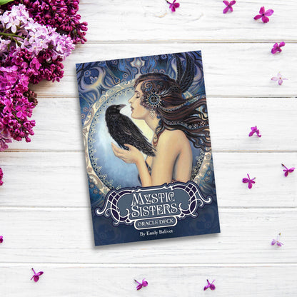 A book titled "Mystic Sisters Oracle Deck" by Emily Balivet is placed on a white wooden surface surrounded by purple flowers. The cover features Emily Balivet artwork of a woman with long hair adorned with intricate designs, holding a black bird, set against a mystical background, evoking feminine wisdom.