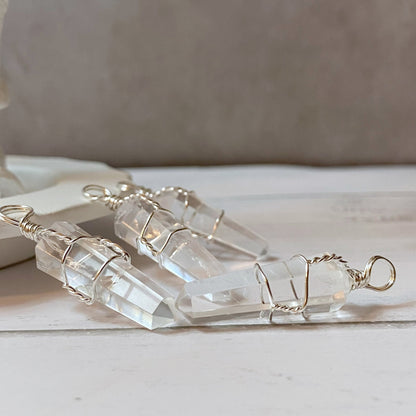 A 1.5" Double Terminated Clear Quartz Point Pendant wrapped in delicate silver wire is suspended from a black cord. This healing pendant, known for fostering spiritual growth, features an elongated crystal with a pointed tip and is attached to a white display stand, set against a plain grey background.