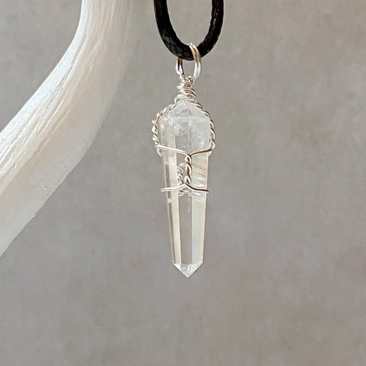 A 1.5" Double Terminated Clear Quartz Point Pendant wrapped in delicate silver wire is suspended from a black cord. This healing pendant, known for fostering spiritual growth, features an elongated crystal with a pointed tip and is attached to a white display stand, set against a plain grey background.