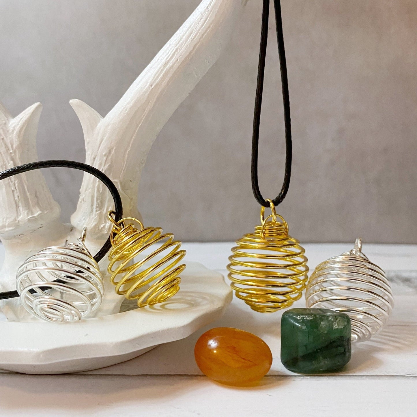 A decorative display of two Empty Spiral Cage Spring Pendants, one in gold and the other in silver, are hung on black cords and draped over a white antler-shaped stand. In front, there are three gemstones—an orange, a green, and a translucent—placed on a textured surface.