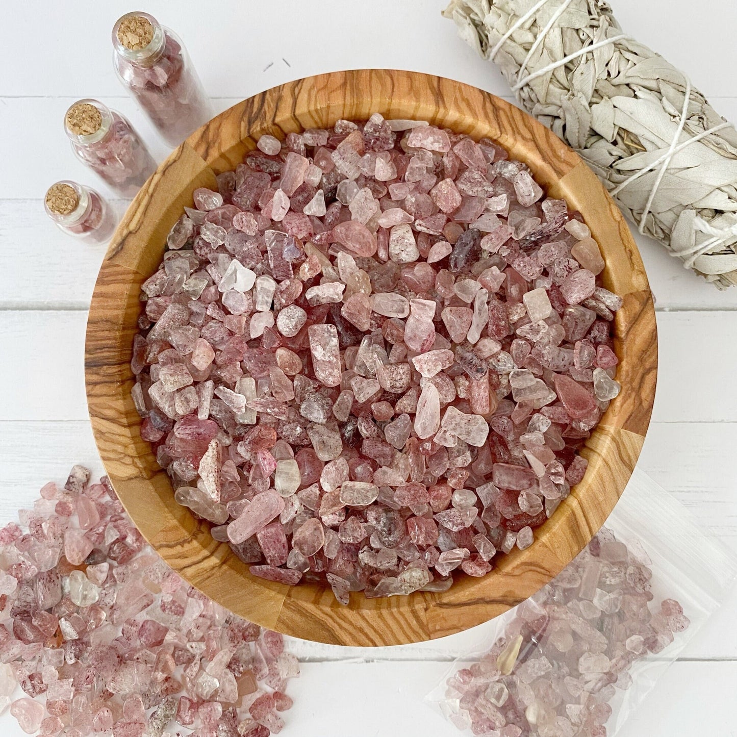 A wooden bowl filled with rose quartz crystals is placed on a white wooden surface. Surrounding the bowl are small glass jars of additional crystals, a bundle of sage, and scattered **Strawberry Quartz Crystal Chips**, hinting at their metaphysical uses.