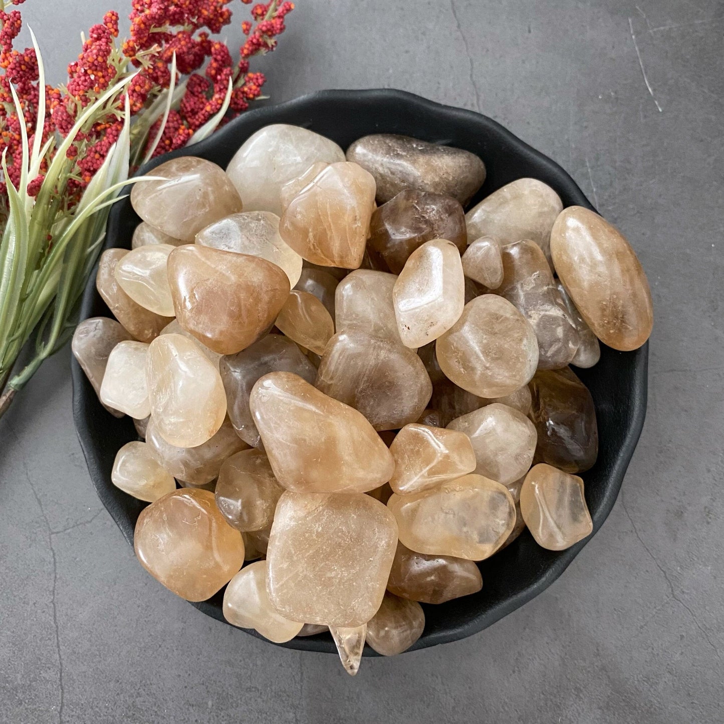 A black bowl filled with various polished, semi-translucent brown and beige gemstones, including a stunning Natural Citrine Tumbled Stones known as a manifestation stone, sits on a gray surface. To the left of the bowl, there is a sprig of red berries and green leaves.