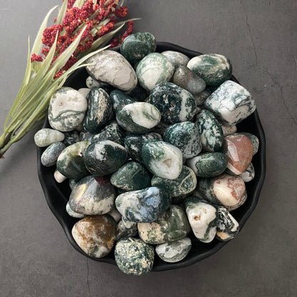 A black bowl filled with smooth, polished Tree Agate Tumbled Stones of varying shades of green, white, and hints of brown. The calming gemstones rest on a dark surface, accompanied by a small bunch of red berries and green foliage, enhancing mental clarity.