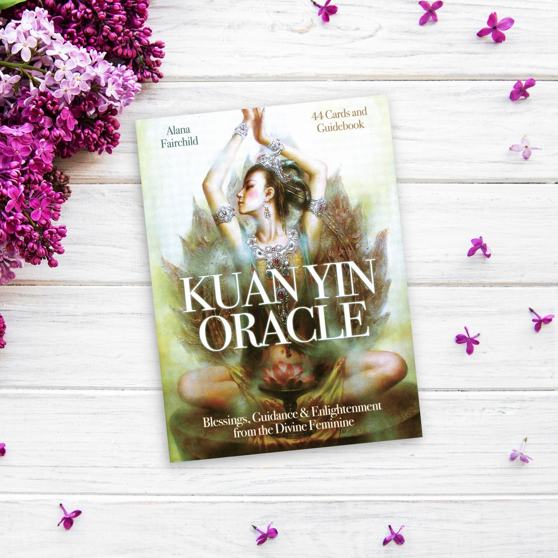 A deck of cards titled "Kuan Yin Oracle" rests on a white wooden surface, surrounded by scattered small purple flowers and sprigs of lilacs. The Kuan Yin Oracle features a serene image of a woman in a meditative pose adorned with intricate jewelry, embodying the essence of the Divine Feminine.