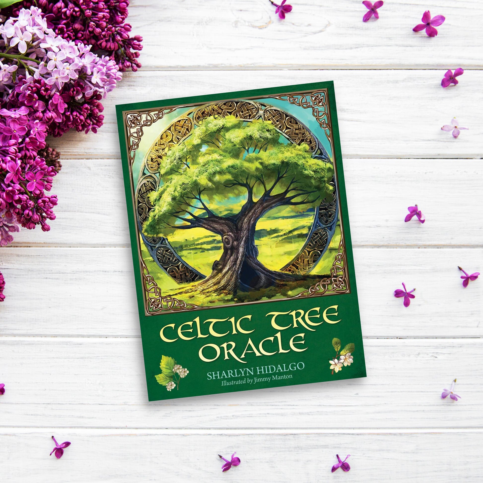 The cover of the "Celtic Tree Oracle" by Sharlyn Hidalgo features an illustration of a large tree set in a circular frame adorned with ancient Celtic designs. Placed on a white wooden surface, the book is surrounded by small purple flowers and lilac blossoms, evoking ancient Celtic wisdom.