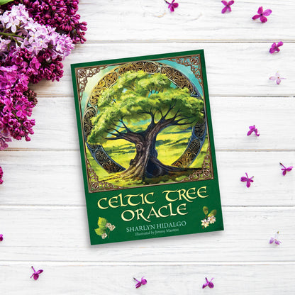 The cover of the "Celtic Tree Oracle" by Sharlyn Hidalgo features an illustration of a large tree set in a circular frame adorned with ancient Celtic designs. Placed on a white wooden surface, the book is surrounded by small purple flowers and lilac blossoms, evoking ancient Celtic wisdom.