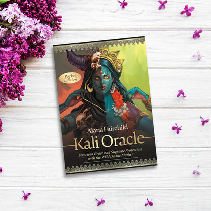 The image shows a book titled "Pocket Kali Oracle" by Alana Fairchild, placed on a white wooden surface adorned with scattered purple flowers. The book cover features an illustration of the goddess Kali, depicted with multiple arms and wearing traditional attire, symbolizing spiritual freedom and radical transformation.